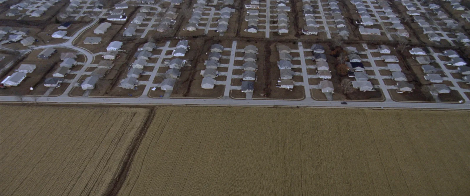 Aerial view in which a dense grid of subdivision rooftops fills the upper right and open ploughed field fills the lower left, with a hard straight edge running diagonally between the two.