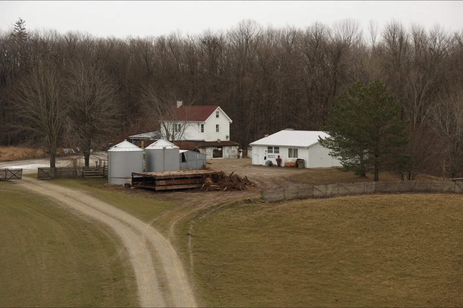 A white two-story farmhouse with a red roof beside a large steel machine shed, two grain bins and a stack of lumber, with a gravel drive forking through winter pasture toward a bare treeline.