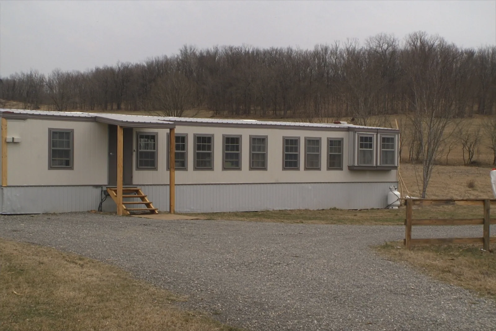 A long single-story manufactured home with a low-pitched roof and a row of identical windows, grey skirting along its base, wooden entry steps, a gravel drive, a propane tank at the far end and winter pasture and bare treeline behind.