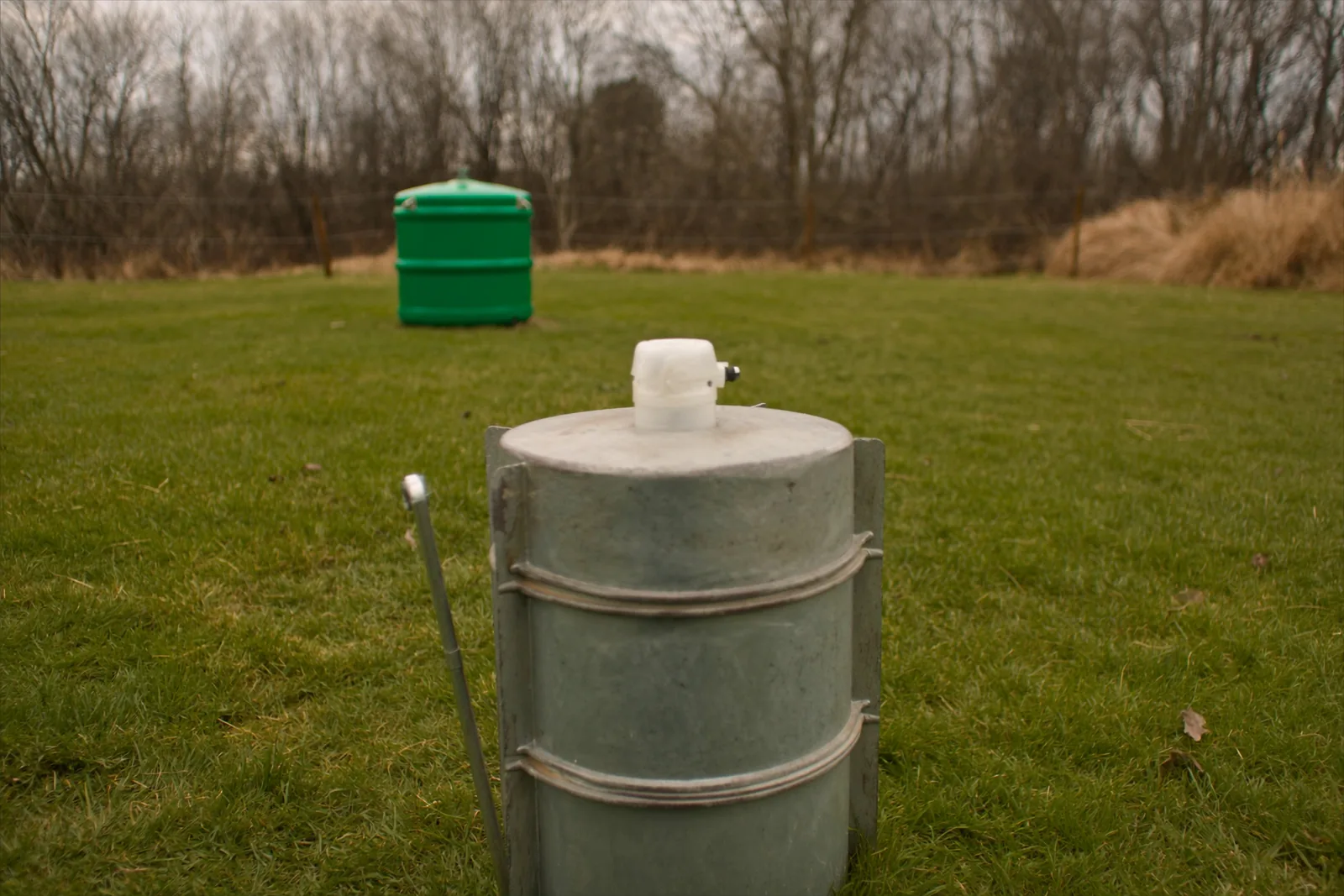 A galvanized steel well casing with a capped pitless adapter standing above mown grass in a rural yard, with a wire fence and a bare treeline behind.